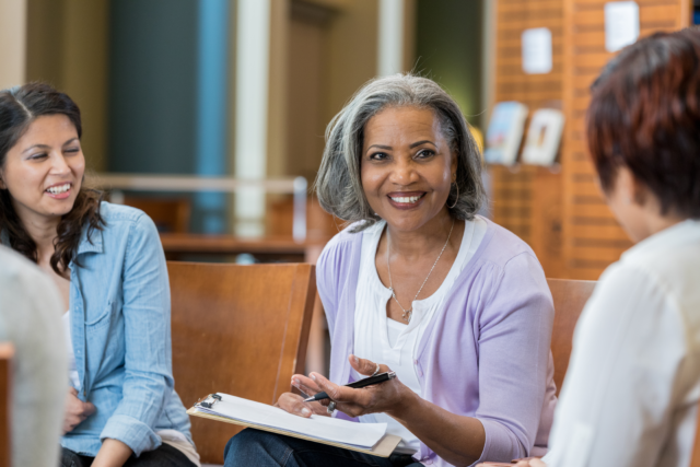 A senior female university professor sits in a circle with her class in the library.  She holds a clipboard as she speaks to the group.